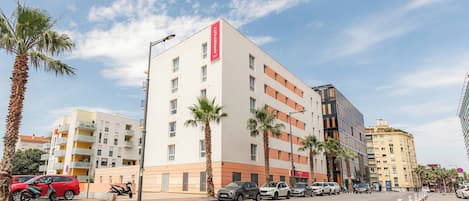 Un hotel de varios pisos con fachada blanca, palmeras y automóviles estacionados en la calle.