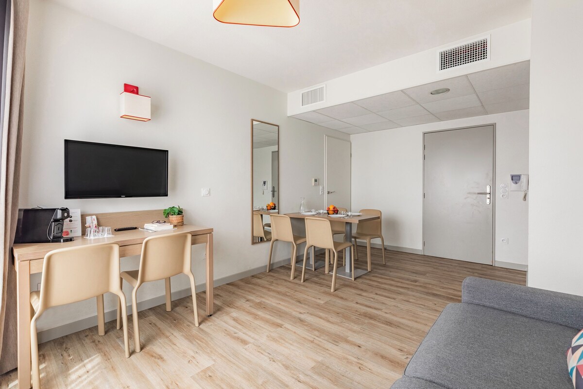 A modern hotel room with a flat-screen TV, a dining area with a table and chairs, a sofa, and a large mirror.