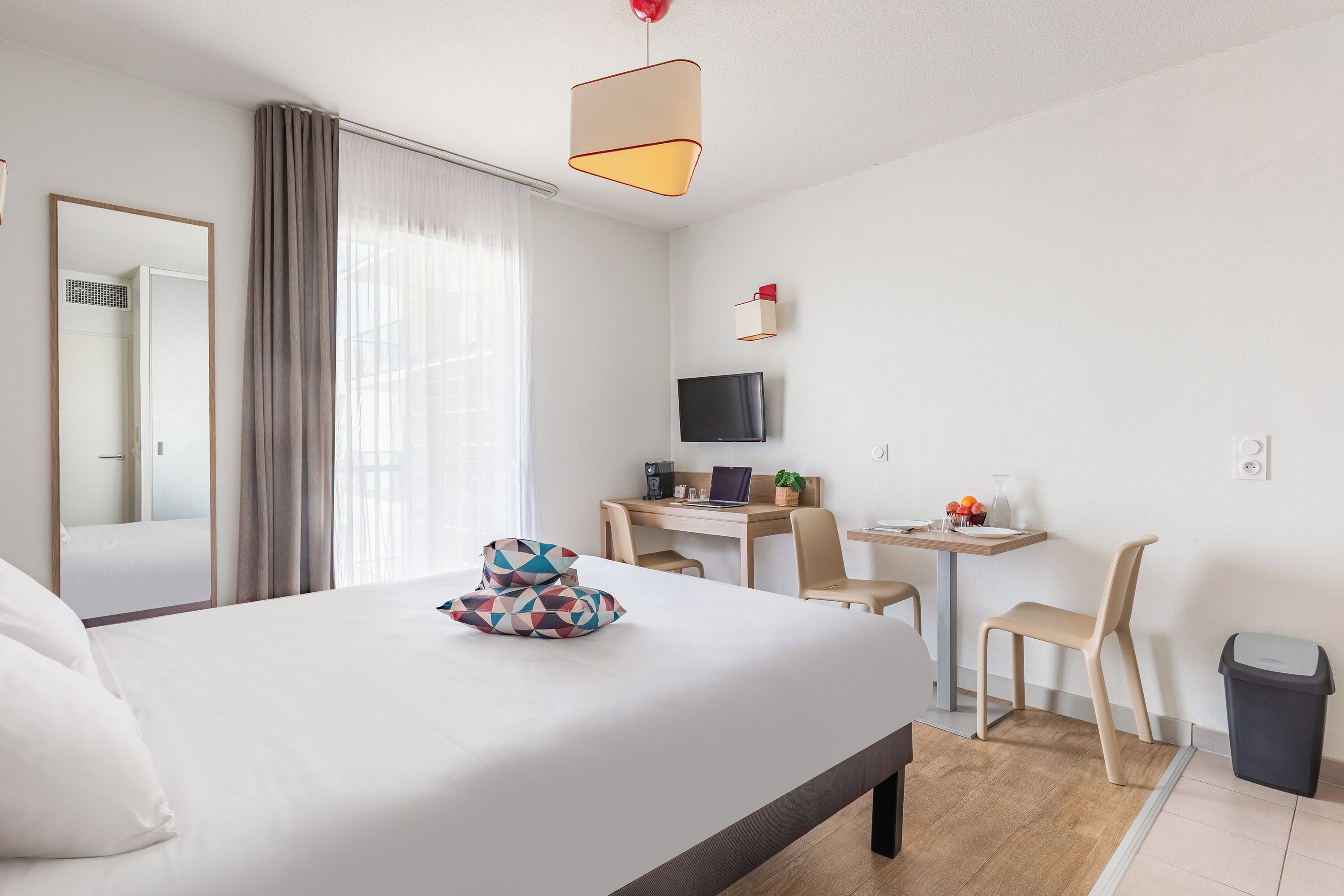 A hotel room with a bed, a desk with a computer, a chair, a TV, and a trash bin.