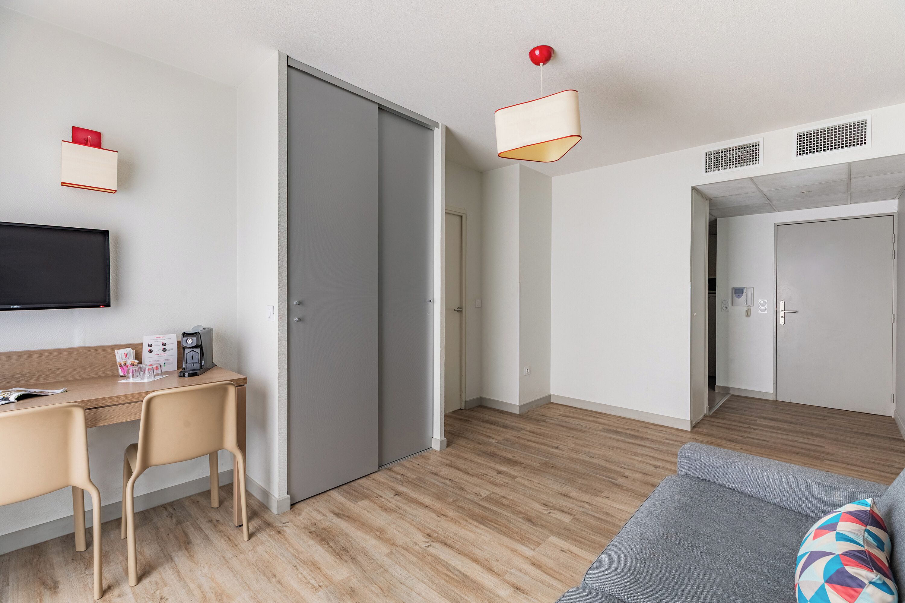 A modern hotel room with a wooden floor, a grey sofa, a desk with a chair, a flat-screen TV, and a sliding door closet.