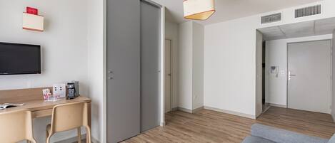 A modern hotel room with a wooden floor, a grey sofa, a desk with a chair, a flat-screen TV, and a sliding door closet.