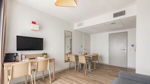 A modern hotel room with a flat-screen TV, a dining area with a table and chairs, a sofa, and a large mirror.