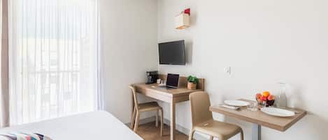 A modern hotel room with a bed, a desk with a laptop, a TV, and a small table with fruits and a vase.