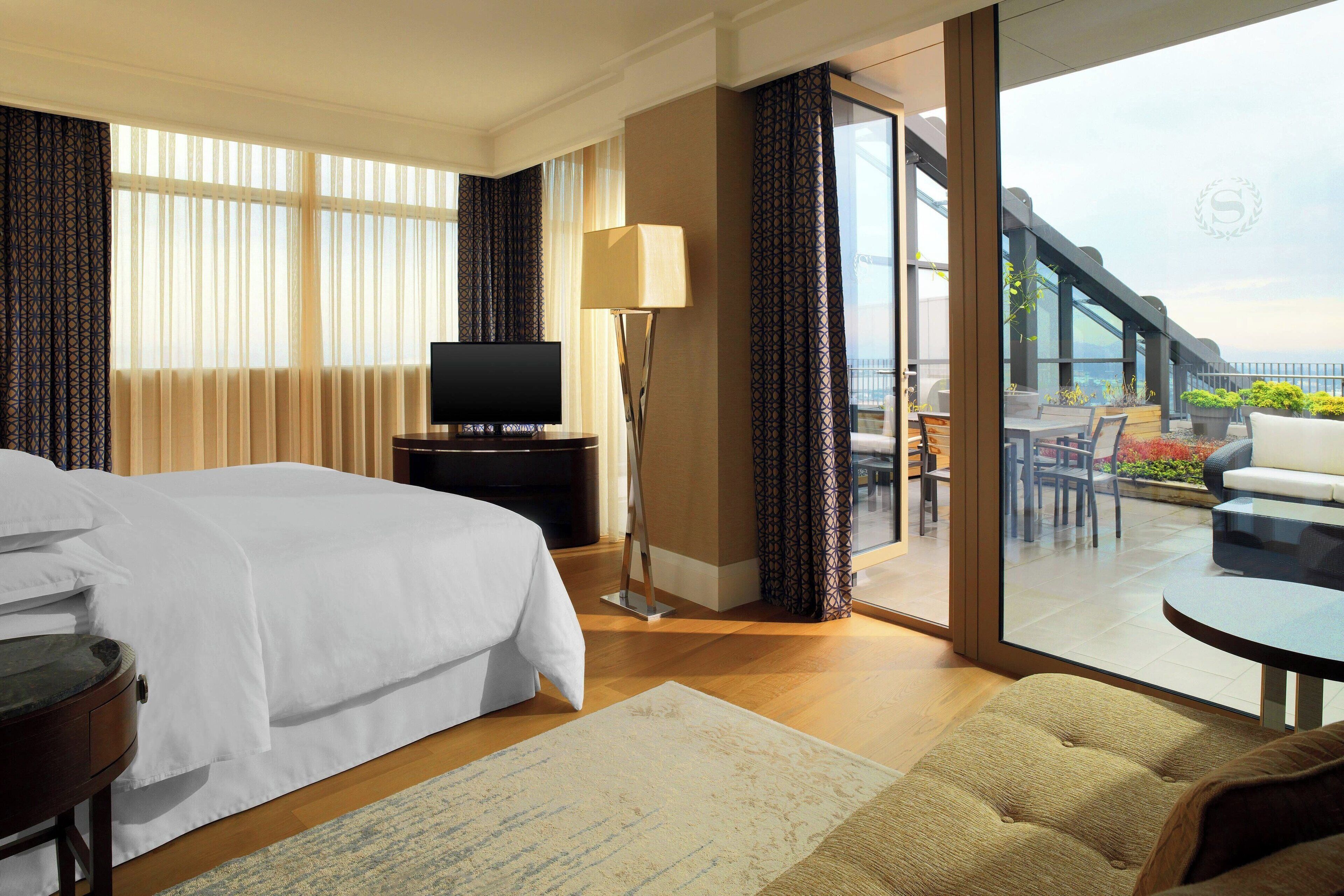 A hotel room with a bed, a TV, a lamp, a sofa, and a balcony with outdoor seating.