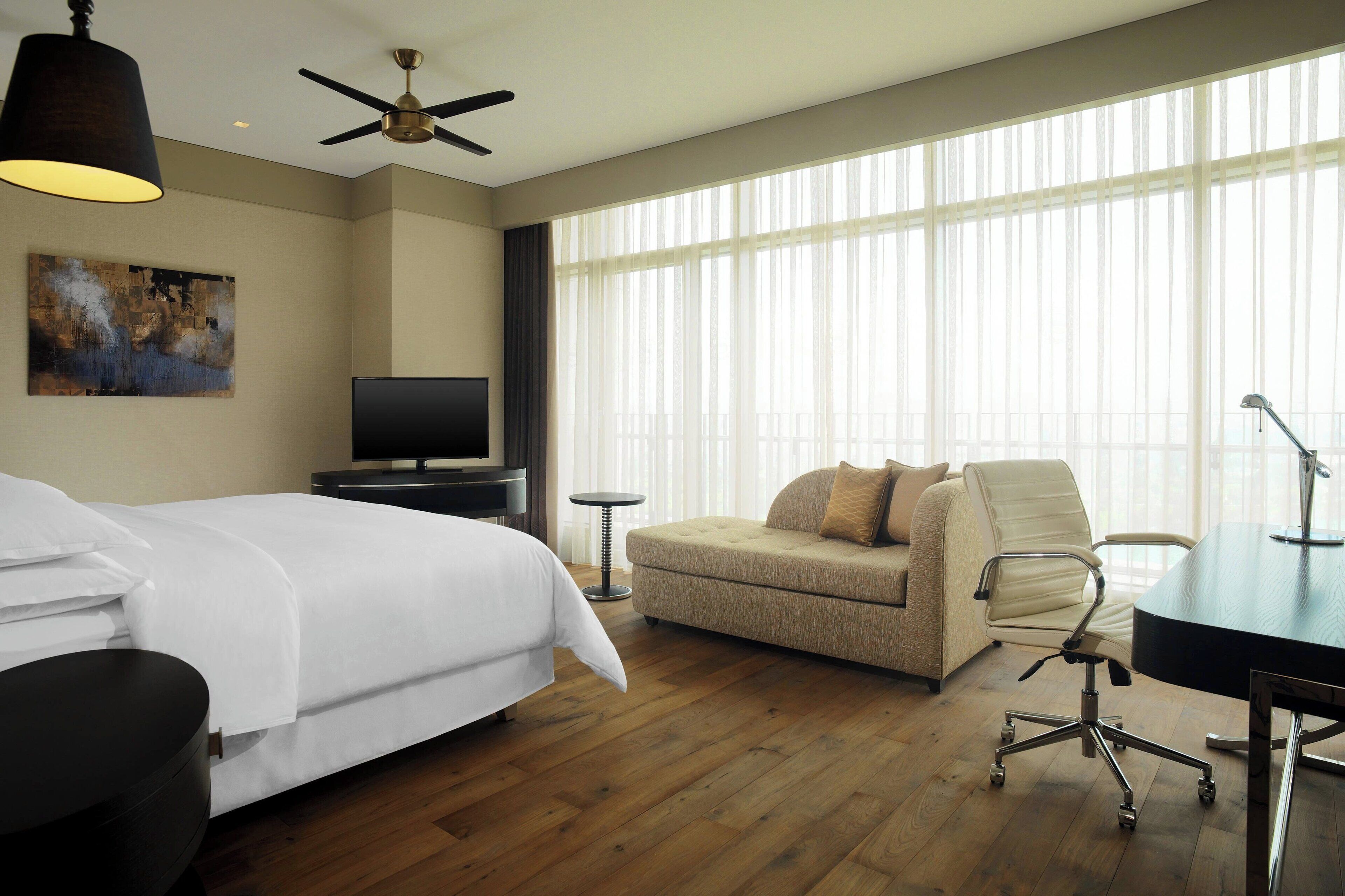 A hotel room with a bed, a sofa, a desk, a TV, and large windows.