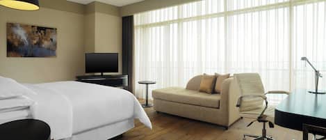 A hotel room with a bed, a sofa, a desk, a TV, and large windows.