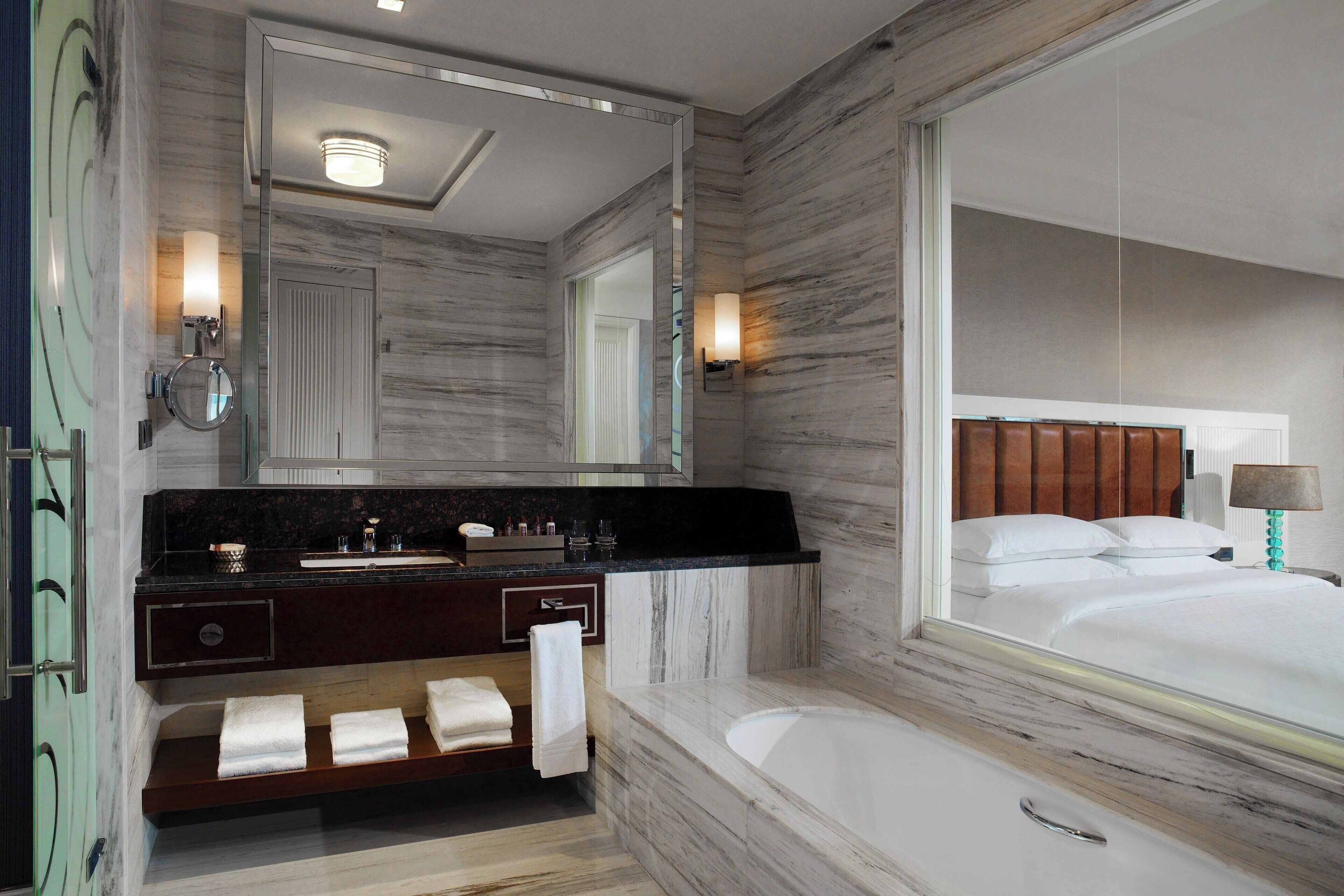 A modern bathroom with a marble vanity, a large mirror, a bathtub, and a bed visible through the mirror.