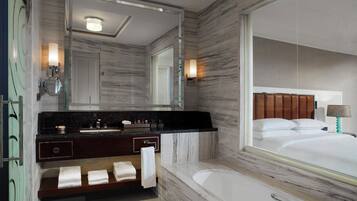 A modern bathroom with a marble vanity, a large mirror, a bathtub, and a bed visible through the mirror.