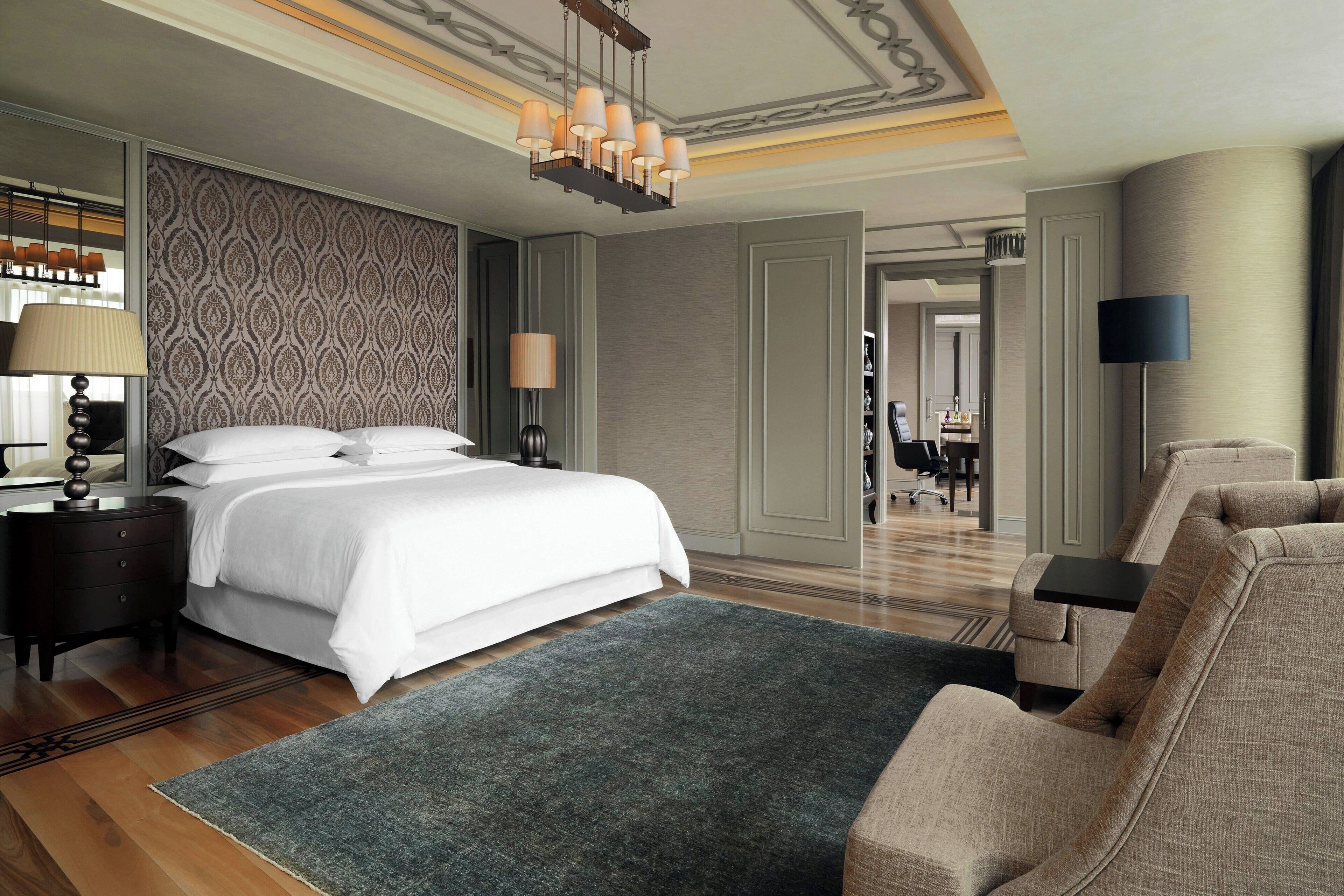 A hotel room with a large bed, a desk, a chair, a lamp, and a patterned wall.
