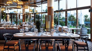 A restaurant with round tables set for dining, large windows offering a view of the outdoors, and hanging pendant lights. - Sheraton Grand Adana (Adana)