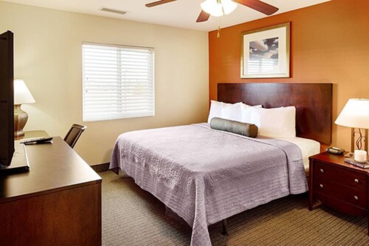 Een hotelkamer met een bed, bureau, stoel, nachtkastje, lamp, plafondventilator en een raam met jaloezieën.