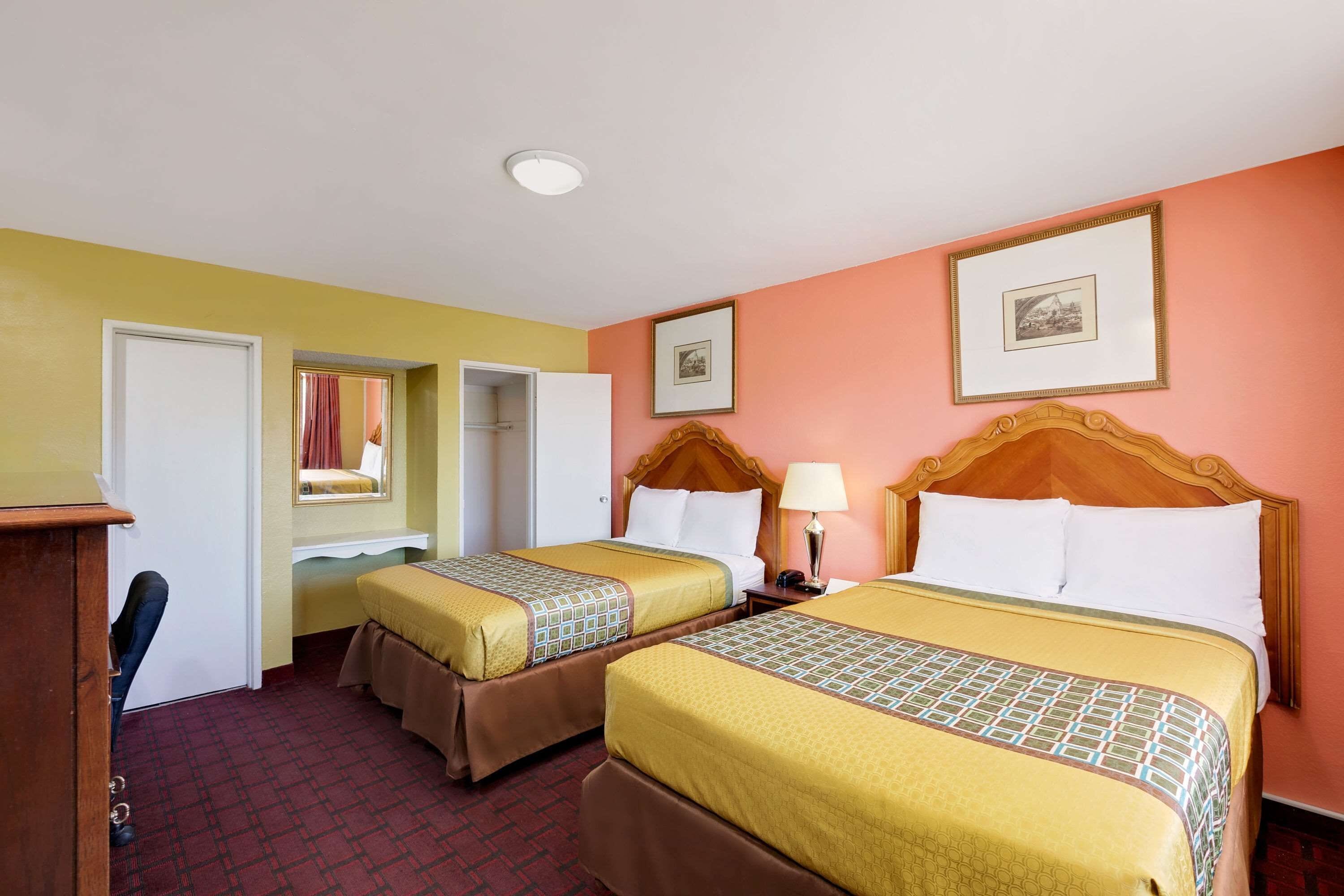 A hotel room with two beds, a wooden headboard, a bedside table with a lamp, and a framed picture on the wall.