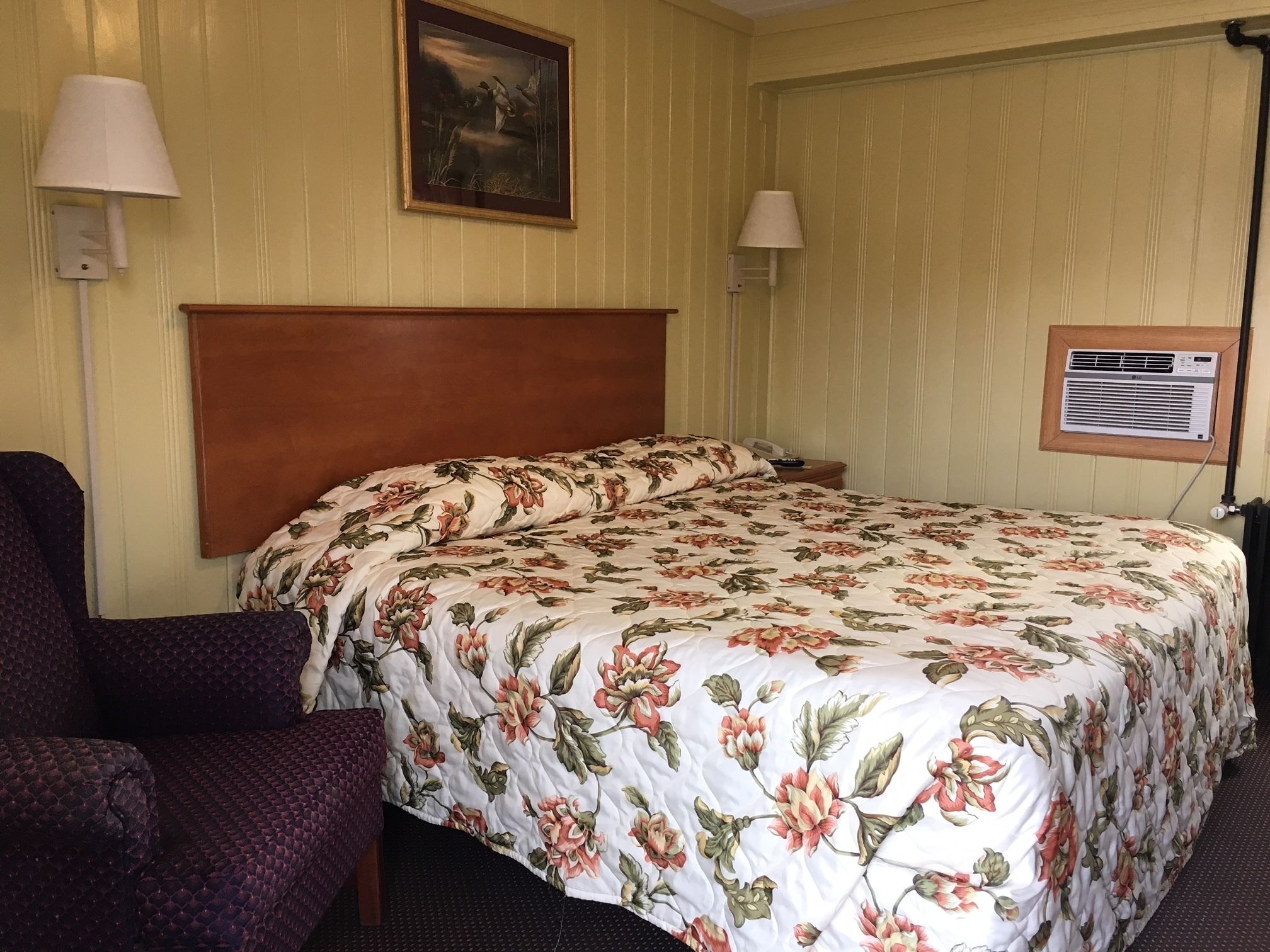 A hotel room with a floral bedspread, a wooden headboard, two bedside lamps, a chair, and a painting on the wall.