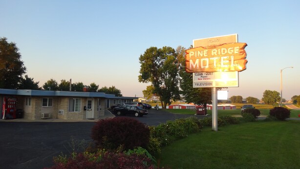 Un panneau indiquant le « Pine Ridge Motel », avec les mentions suivantes : chambres propres, téléphones, nouveaux propriétaires et télévision.