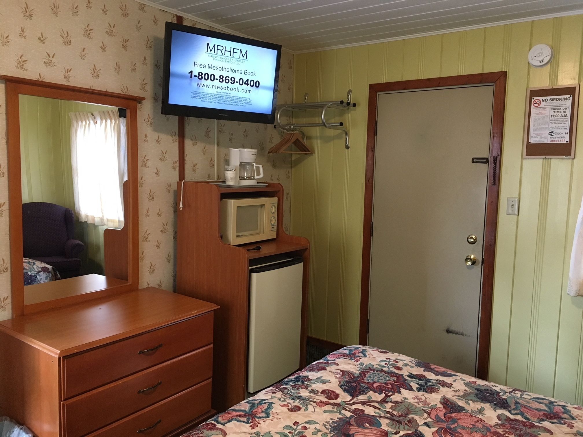 Habitación de hotel con un aparador de madera, una cama con colcha estampada, un televisor sobre el aparador mostrando un anuncio de libros, y una puerta con un cartel de 'No Fumar'.