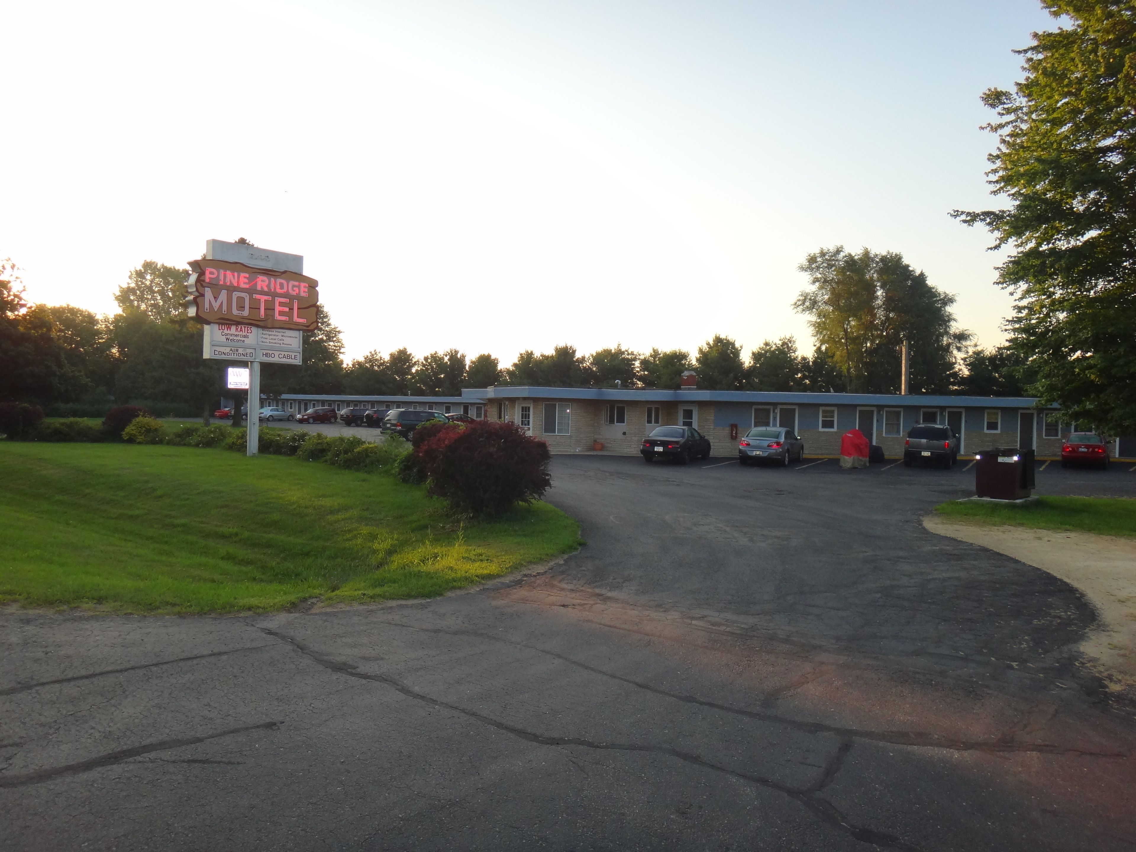 Pine Ridge Motel met een parkeerplaats, een gebouw met een blauw dak en een bord met daop advertentie voor lage prijzen en zakelijke gasten.