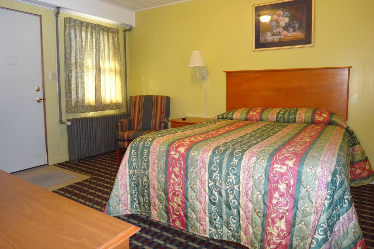 A hotel room with a bed, a chair, a desk, a lamp, and a painting on the wall.