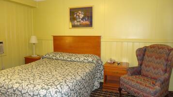 Quarto de hotel com uma cama, uma cadeira, uma mesa de cabeceira, um abajur e um quadro na parede.