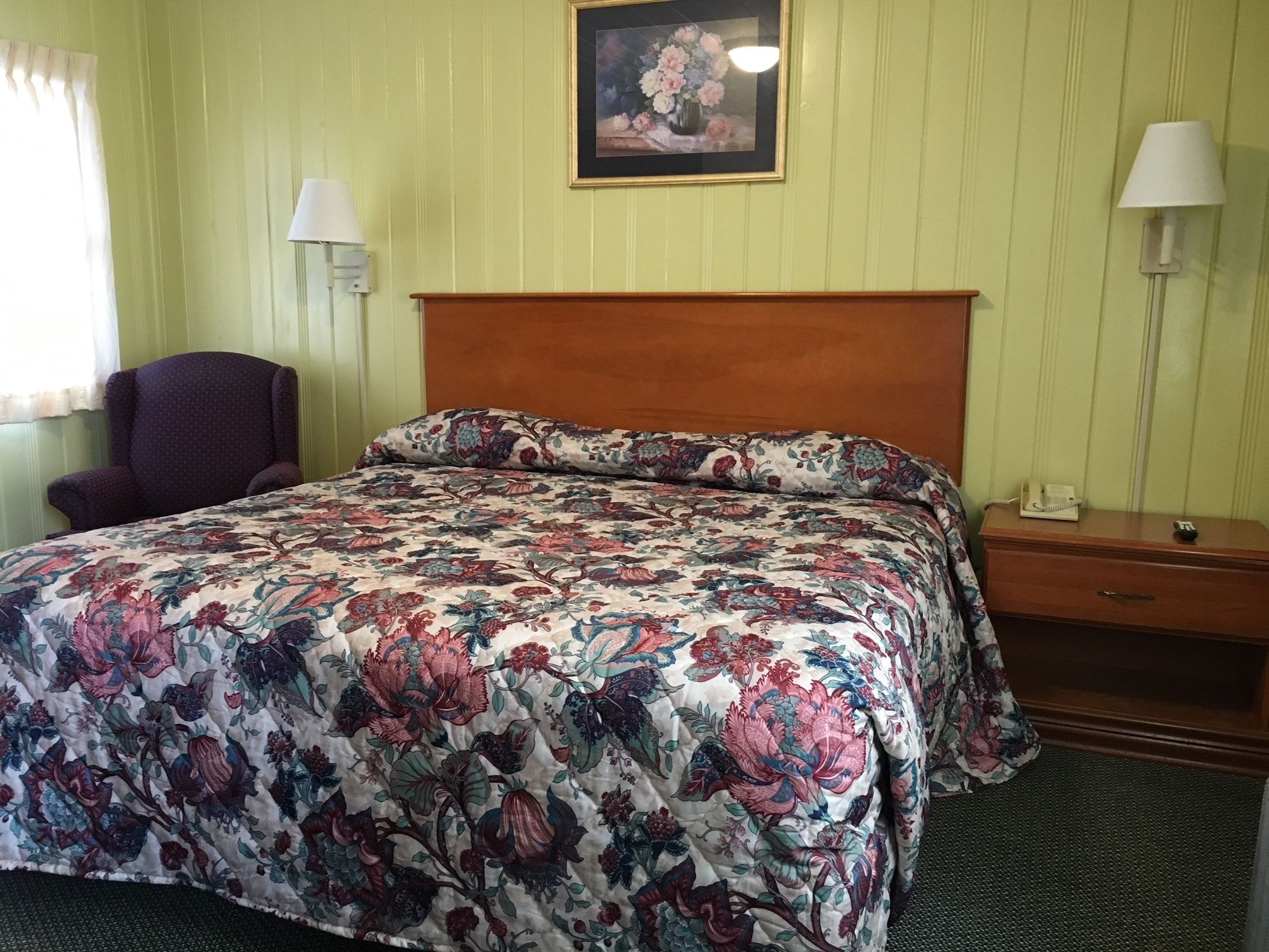A hotel room with a floral-patterned bedspread, a wooden headboard, a chair, a nightstand with a lamp, and a framed picture on the wall.