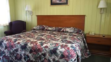 A hotel room with a floral-patterned bedspread, a wooden headboard, a chair, a nightstand with a lamp, and a framed picture on the wall.