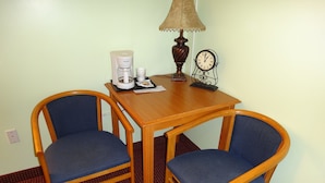 Une petite table en bois, accompagnée de deux chaises bleues à coussin, d’une machine à café, d’une lampe et d’une horloge murale.