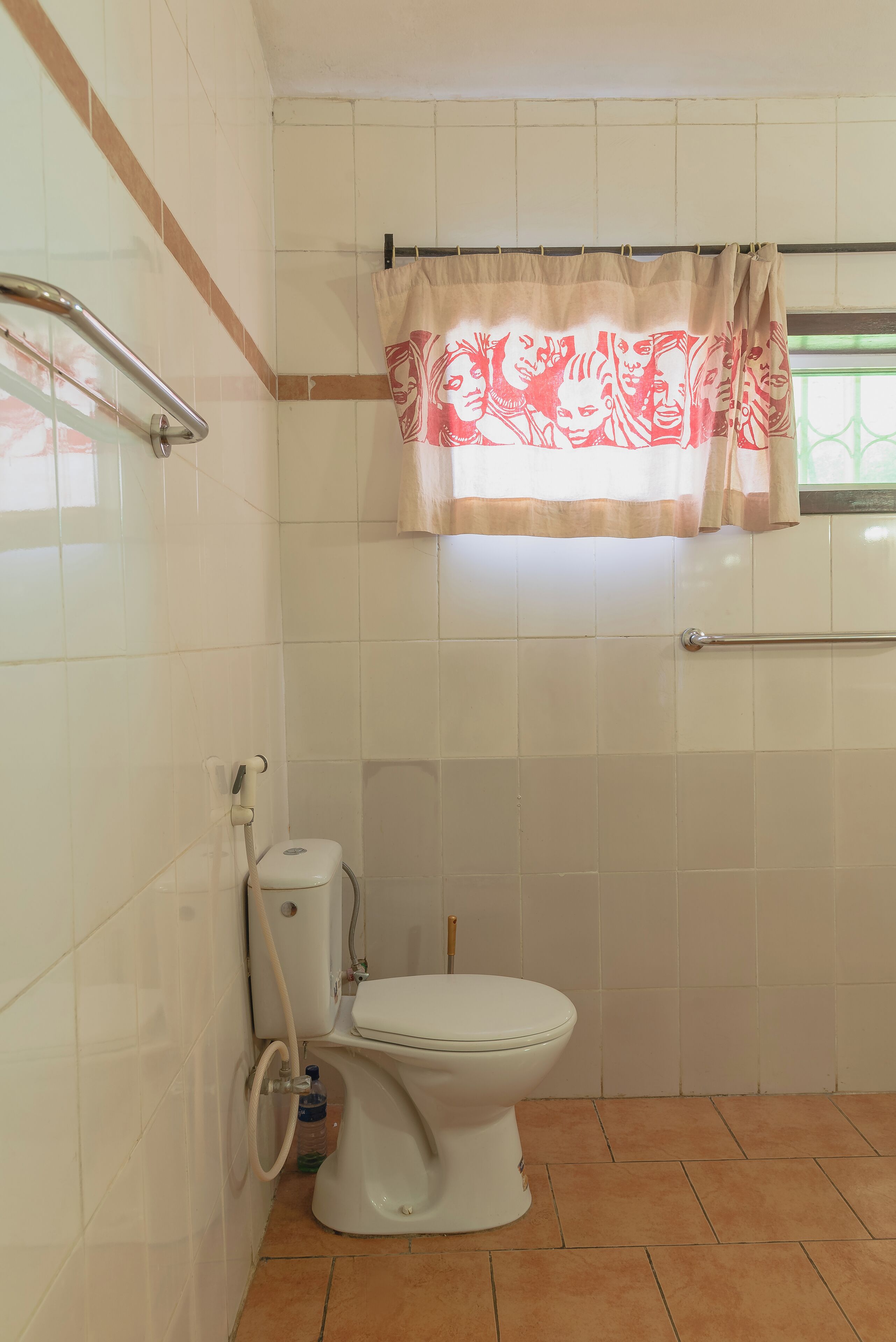 A bathroom with a toilet, a handrail, and a window with a curtain.