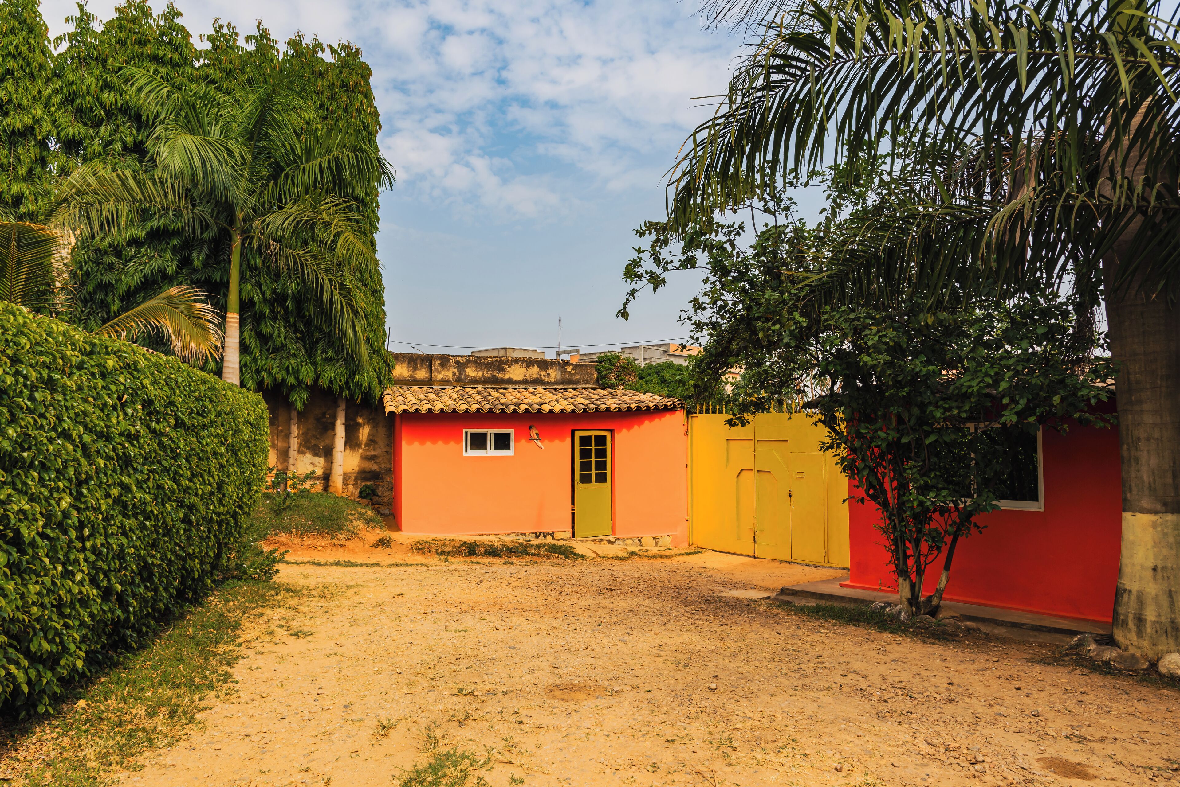 A small, single-story building with a red and yellow facade, surrounded by greenery and a dirt path.