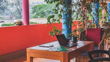 A wooden table with a laptop, notebook, and pen on a balcony with red walls and potted plants.