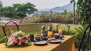 A table set with breakfast items, including fruits, beverages, and a floral centerpiece, overlooking a garden with a mountain view.