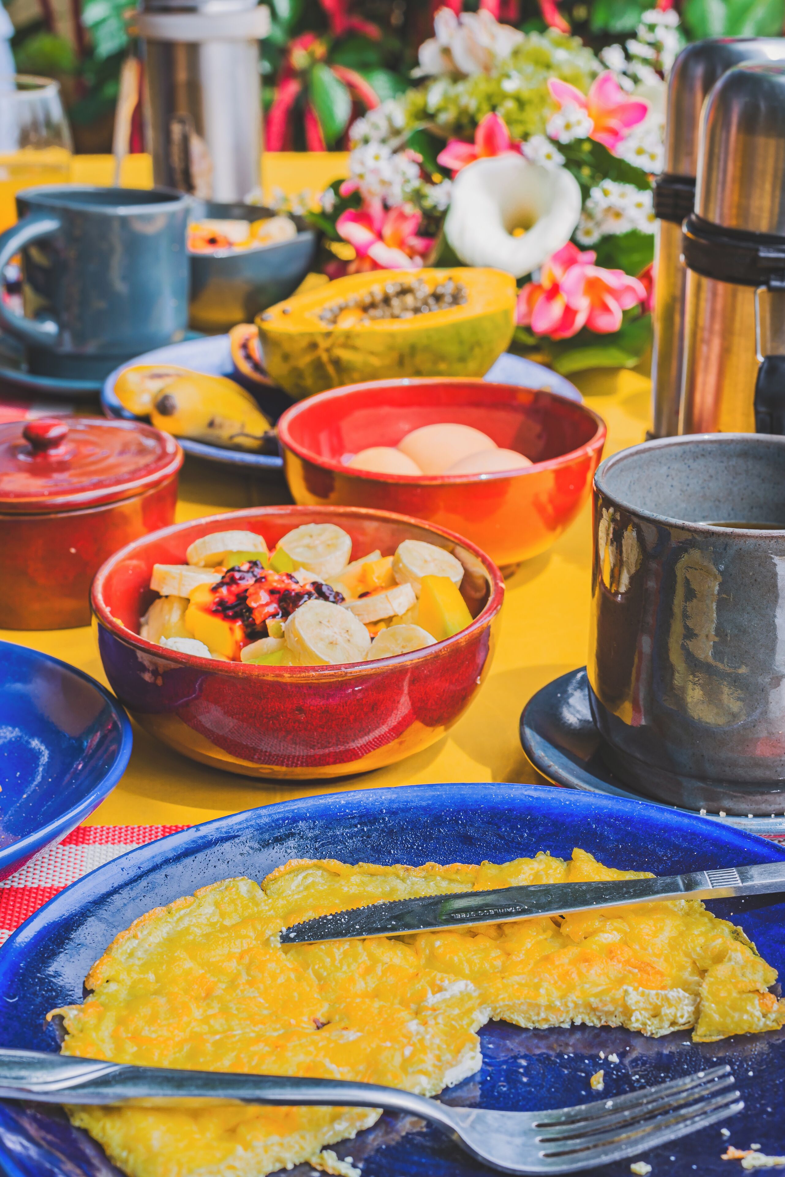 Une table sur laquelle on trouve différents bols remplis de fruits, une assiette avec un plat jaune, peut-être une quiche, et une tasse en métal.