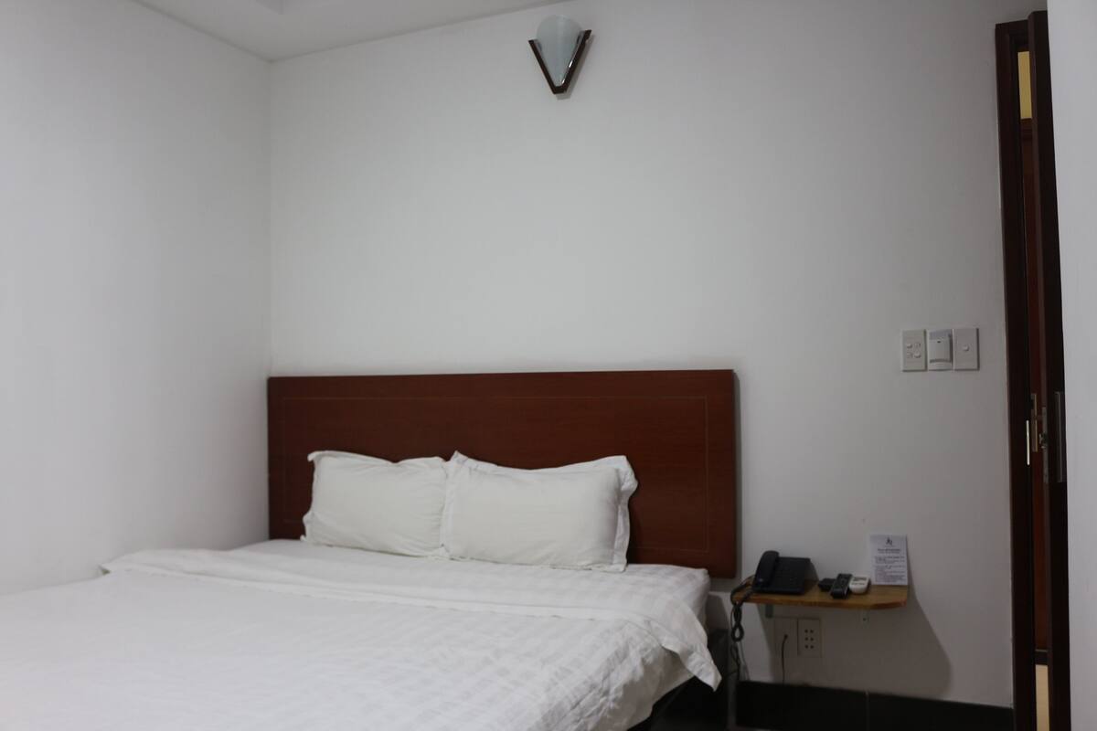 Une chambre d’hôtel avec un lit simple, une tête de lit en bois, une table de chevet avec un téléphone et un panneau, et un luminaire fixé au mur.