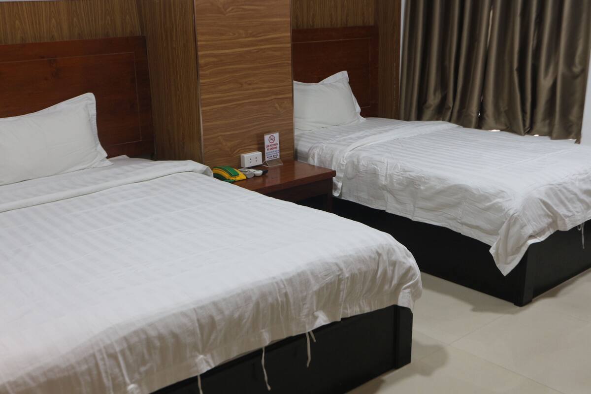 Une chambre d’hôtel avec deux lits, des têtes de lit en bois, une table de chevet avec un téléphone et un panneau, et une fenêtre avec des rideaux.