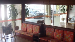 A seating area with floral-patterned cushions, a potted plant, and a view of parked cars outside. - Hosaga Biz Hotel (KELANTAN)