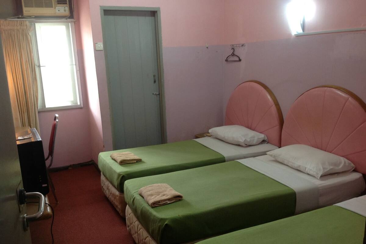 Een kamer met twee eenpersoonsbedden, elk met een groene spreidekker en een wit kussen. Er is een deur met een lichtschakelaar erboven, en een klein tafeltje met een rood voorwerp erop.