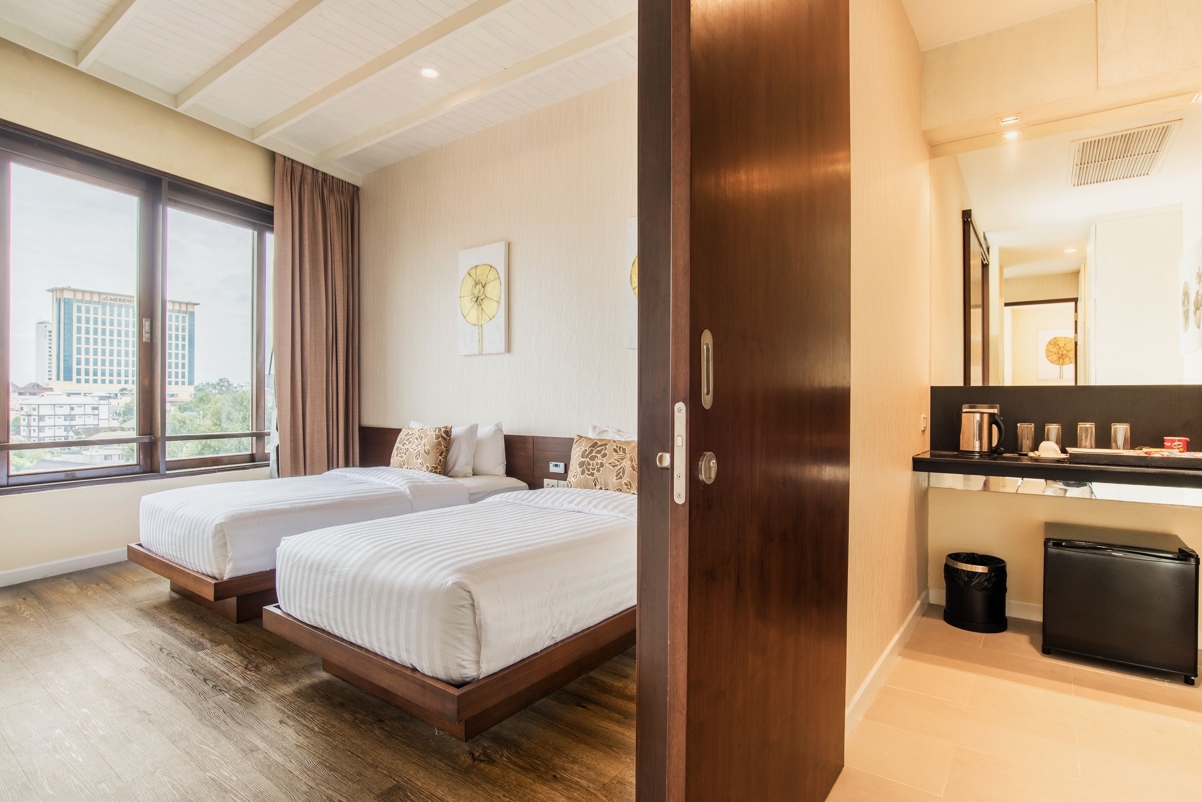 A hotel room with two beds, a wooden floor, a large window, a small kitchen area, and a trash bin.
