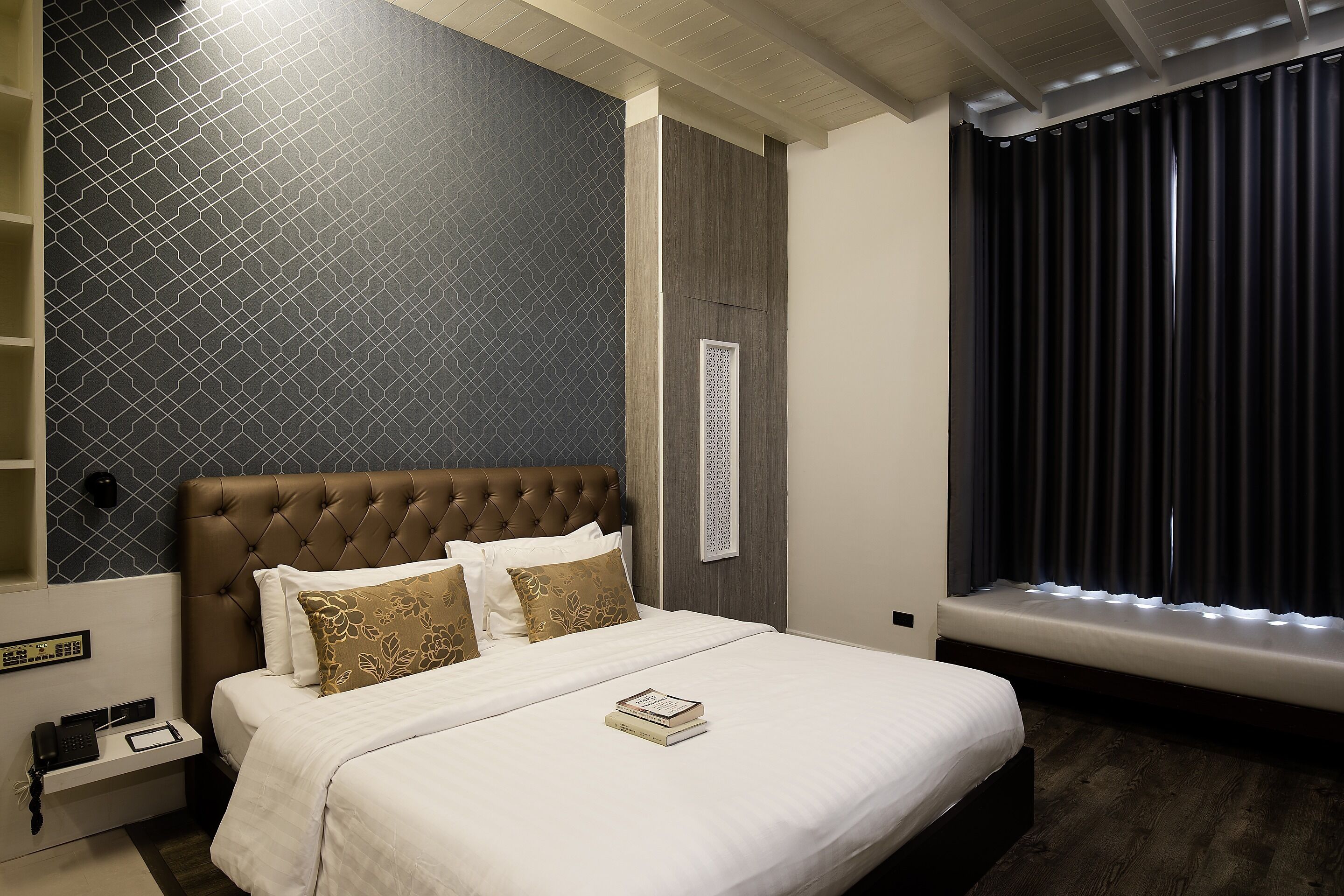 A hotel room with a large bed, a bedside table with a phone and a book, a window with curtains, and a wall with a geometric pattern.