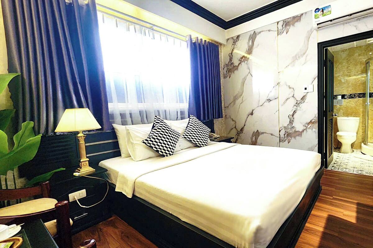 A hotel room with a bed, a nightstand, a lamp, a large window with curtains, a marble wall, and a bathroom visible through an open door.