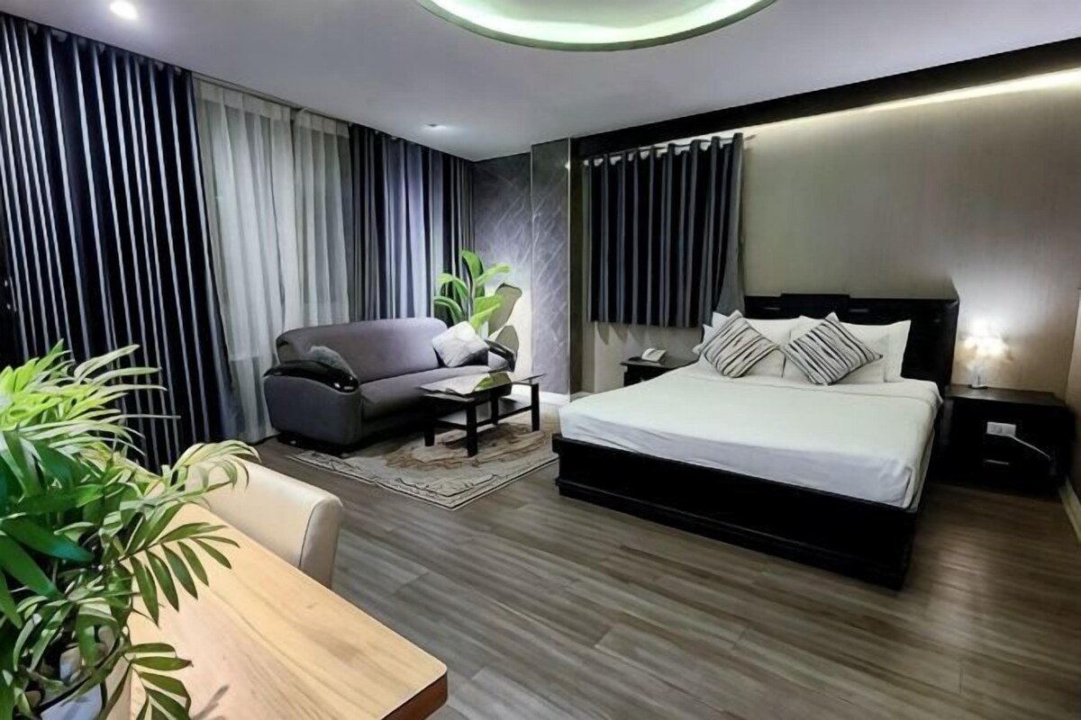 A modern hotel room with a large bed, a sofa, a dining table, and a potted plant.