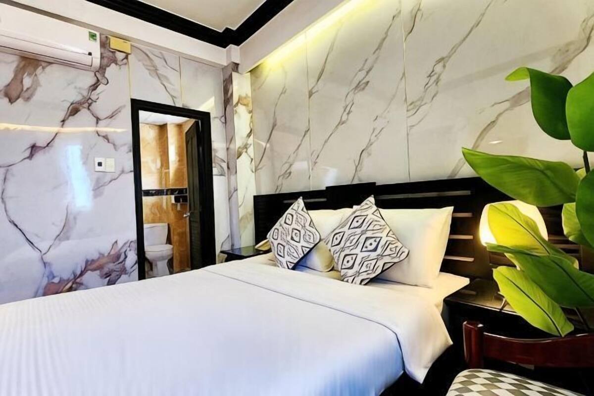 A hotel room with a marble-patterned wall, a bed with patterned pillows, a nightstand with a lamp, and a large green plant.