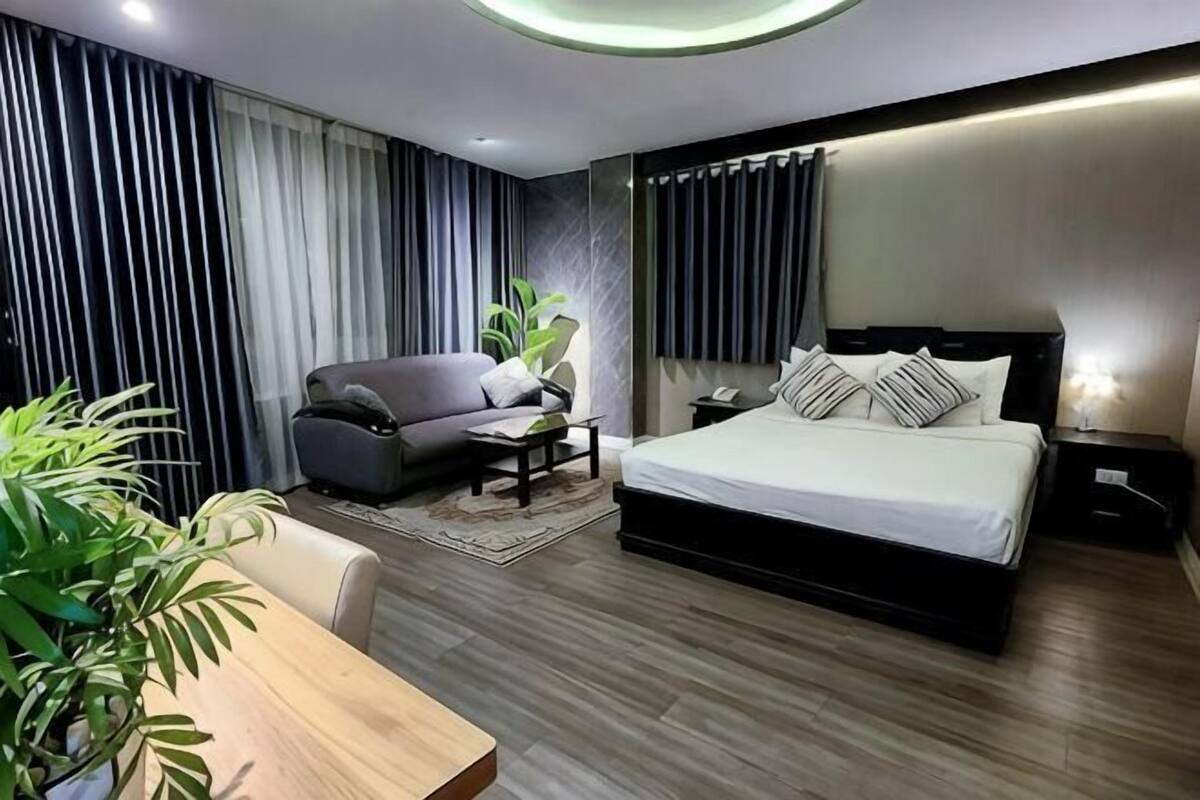 A modern hotel room with a large bed, a sofa, a dining table, and a potted plant.