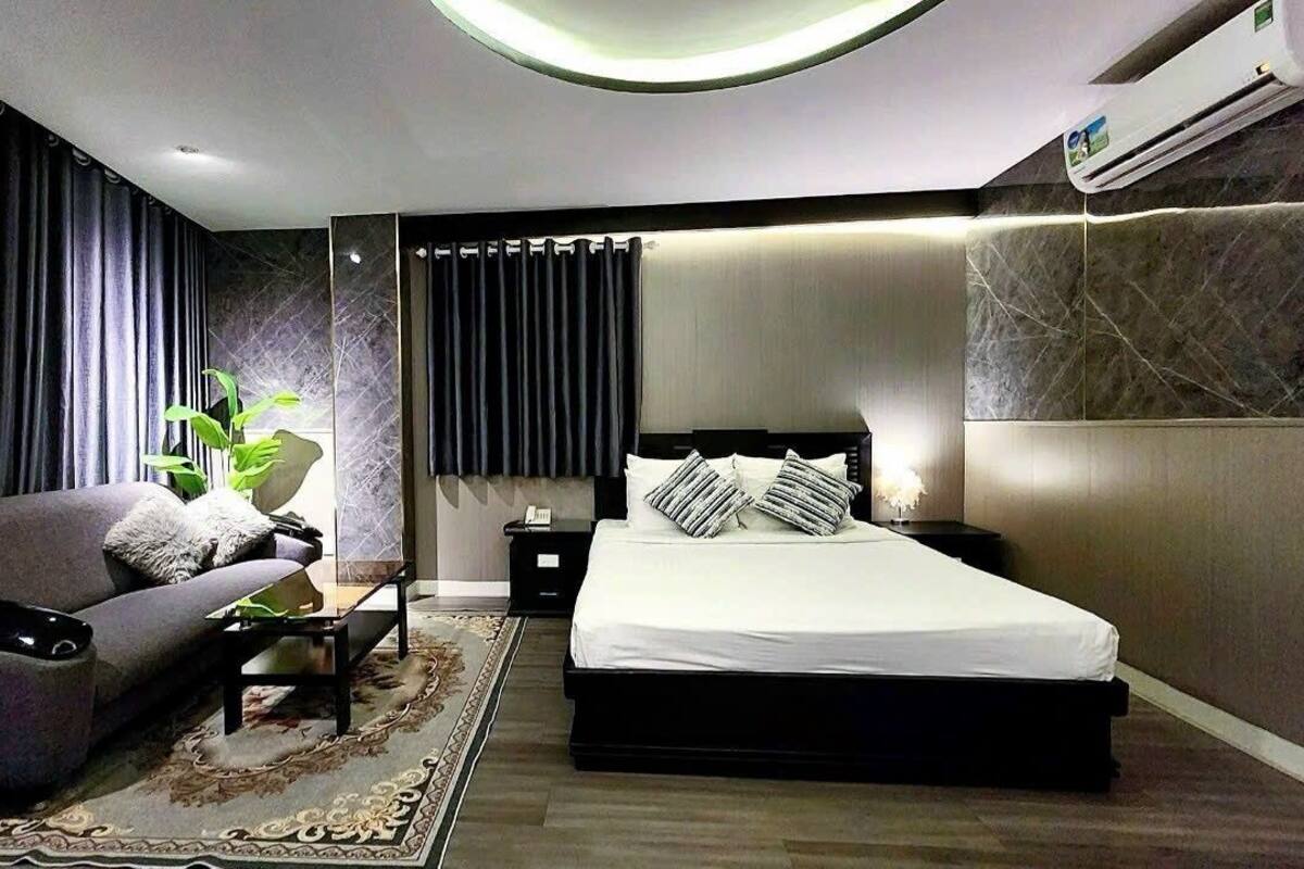 A modern hotel room with a bed, a sofa, a nightstand, and a potted plant.