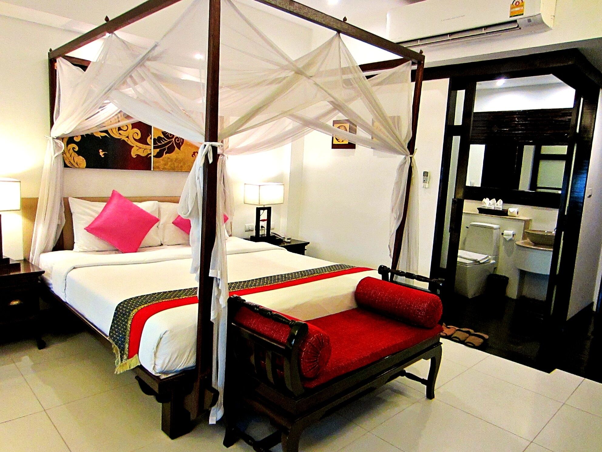 A four-poster bed with a canopy, a red bench, a nightstand, and a bedside table with a lamp.