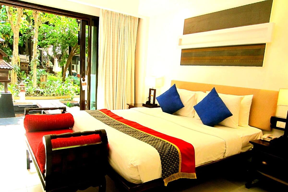 A hotel room with a bed, a desk, and a chair. There is a window with curtains and a view of greenery outside.