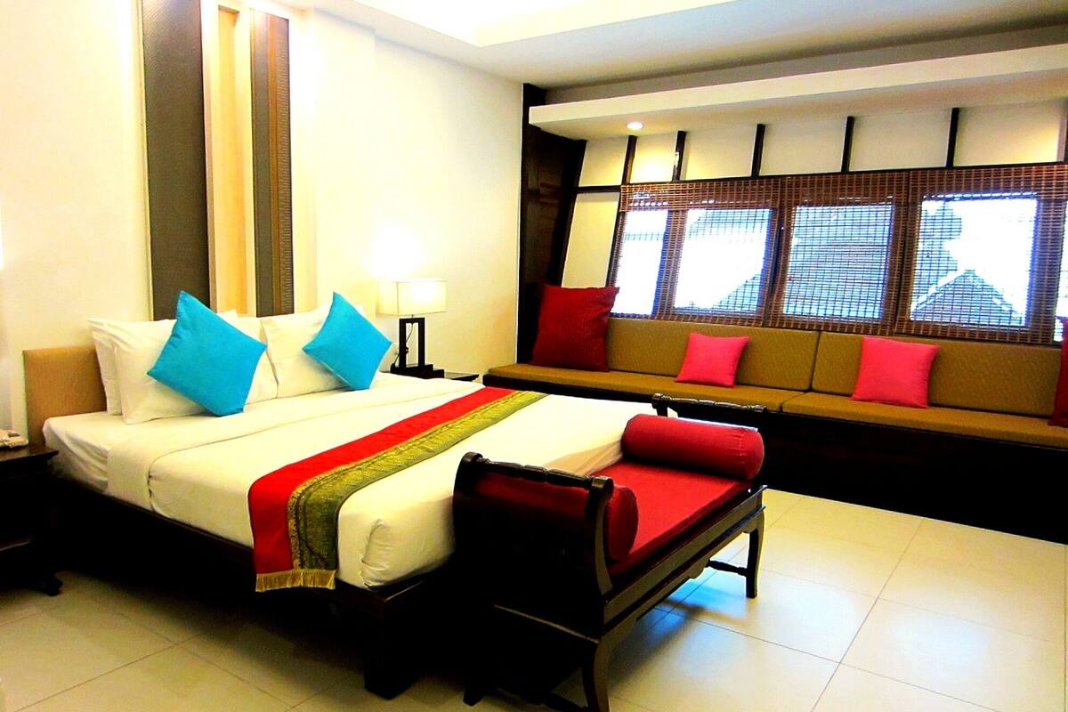 A hotel room with a bed, a sofa, a nightstand, and a window with blinds.