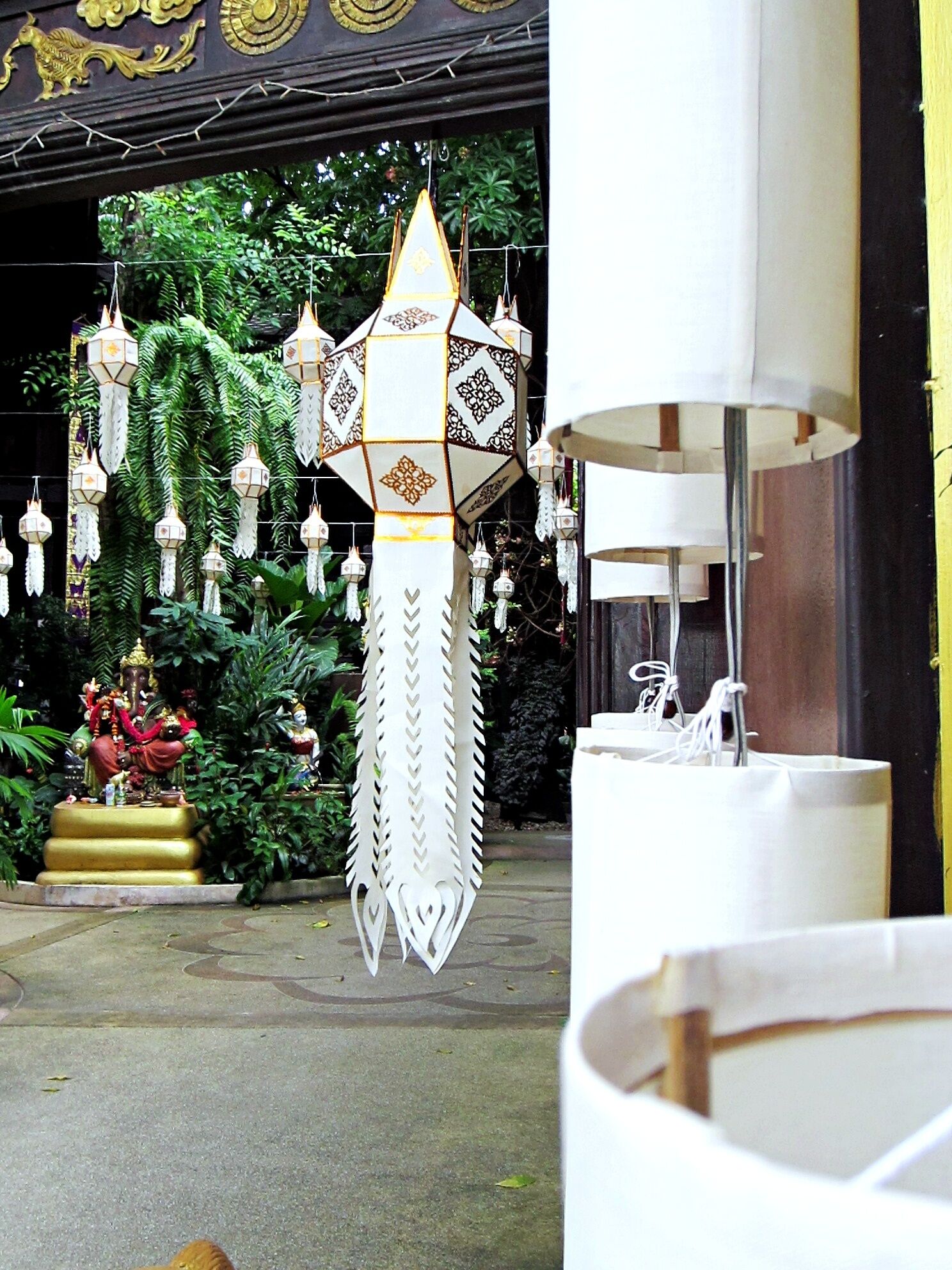 A traditional hanging lamp with a geometric design, a white lamp with a cylindrical shade, and a golden statue of a deity.