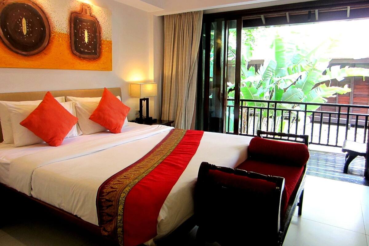 A hotel room with a large bed, red and white bedding, a red and gold bedspread, and a view of greenery through the balcony doors.