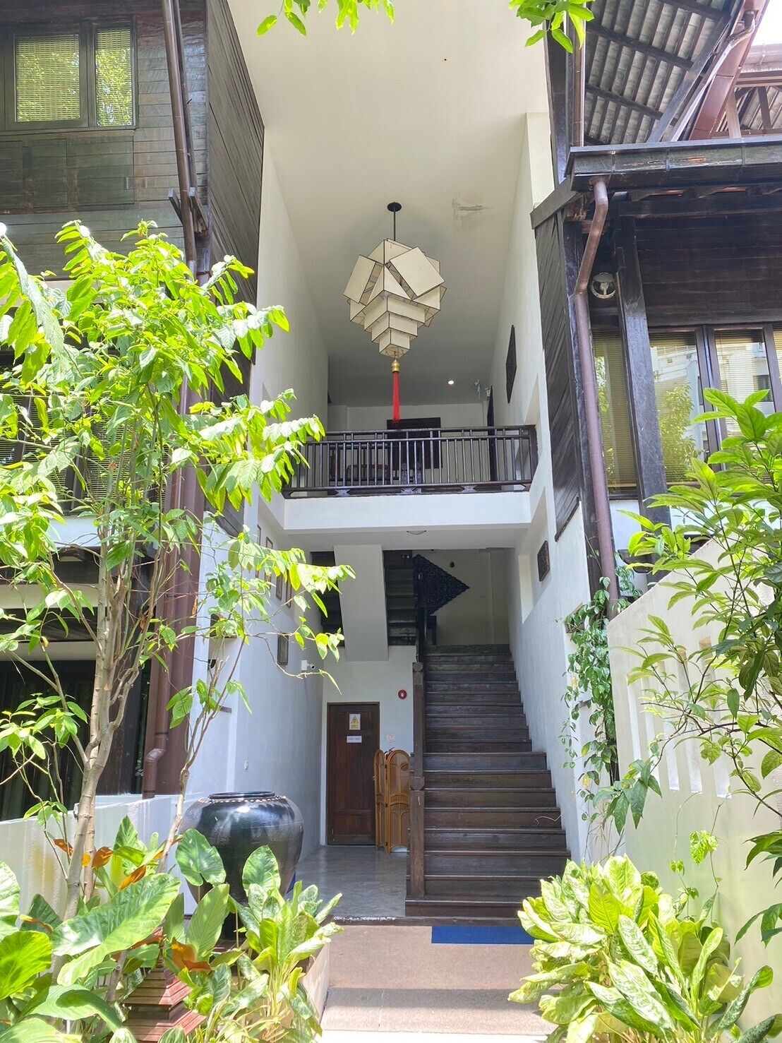 A two-story building with a staircase leading to the upper floor, surrounded by greenery and a large black pot.