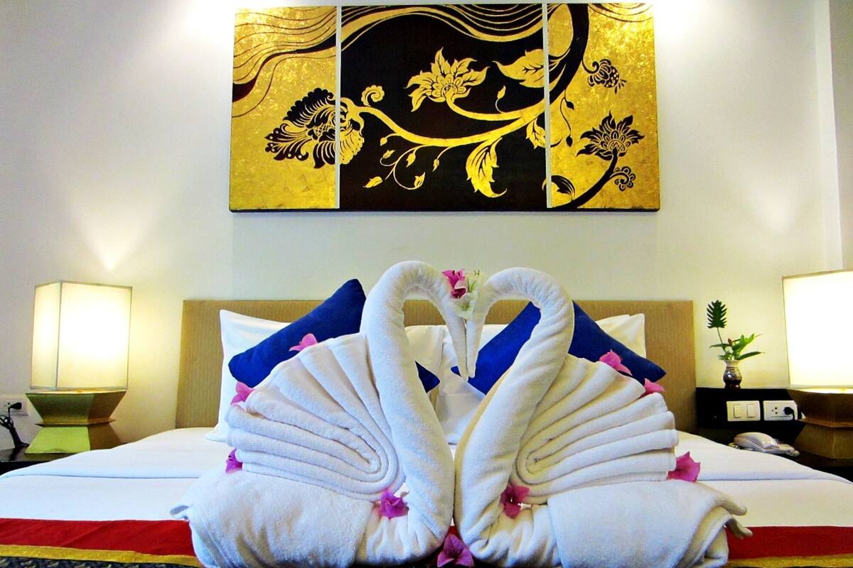 A hotel room with a bed, pillows, a lamp, a wall art piece, and a vase with a plant.