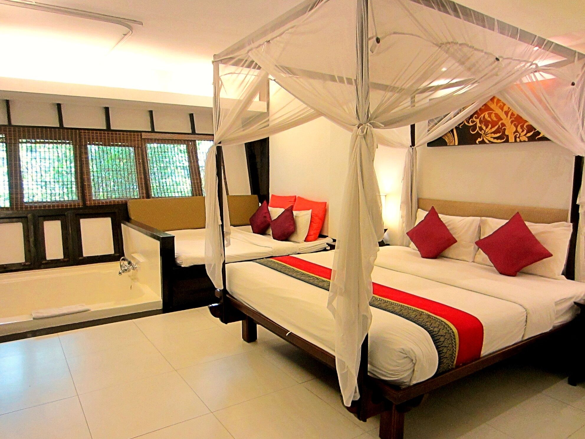 A four-poster bed with white bedding and red pillows, a bathtub, and a window with a view of greenery.