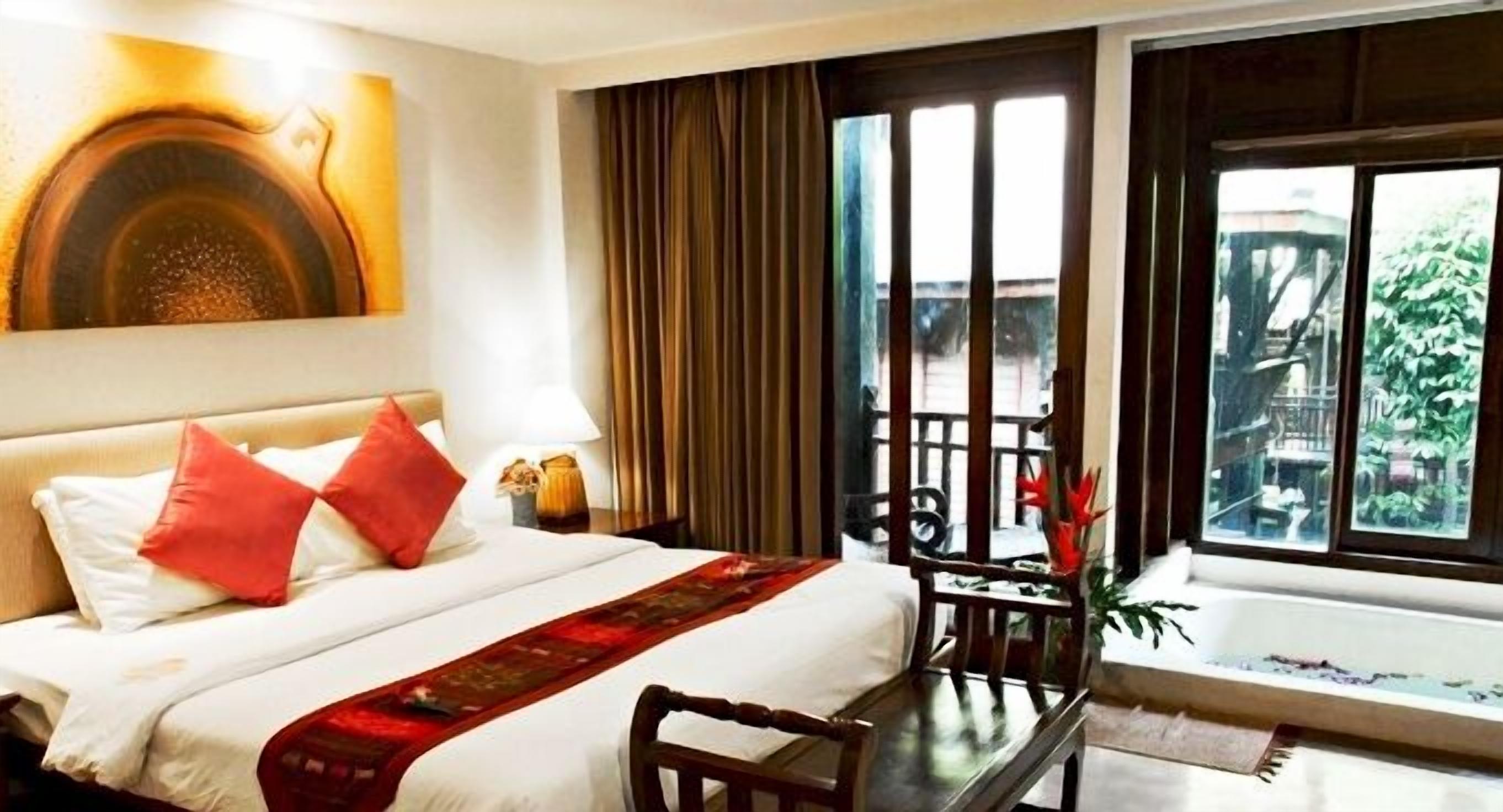 A hotel room with a bed, a desk, a chair, a window with a view of greenery, and a painting on the wall.